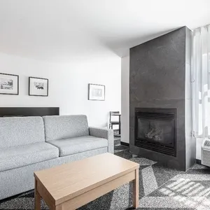 Salon élégant au Doubletree par Hilton Québec Resort, catégorie Salle de réception. La pièce présente un canapé moderne gris clair face à une table en bois claire, avec un élégant foyer encastré dans un mur sombre. Trois cadres d'art en noir et blanc ornent le mur, tandis que de grandes fenêtres laissent entrer une abondance de lumière naturelle à travers des rideaux blancs transparents. Le sol est recouvert d'un tapis à motifs géométriques, ajoutant une touche contemporaine à l'ambiance accueillante, parfaite pour des réceptions de mariage élégantes.