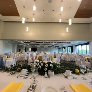La salle de réception du Doubletree par Hilton Québec Resort est élégamment aménagée pour un mariage. Des tables rondes sont magnifiquement dressées avec des nappes blanches et des serviettes jaunes, ajoutant une touche de couleur vibrante. Chaque table est ornée de compositions florales centrées, composées de fleurs blanches et jaunes accompagnées de feuillage vert, créant un cadre sophistiqué. Des chaises couvertes de housses blanches sont disposées autour des tables, offrant une ambiance chic et accueillante. Les lumières suspendues au plafond ajoutent une touche moderne et chaleureuse à cet espace spacieux et lumineux, parfait pour célébrer un mariage inoubliable.