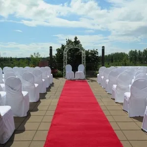 Cette image montre l'aménagement de la salle de réception au Doubletree par Hilton Québec Resort, une catégorie de lieu de mariage. L'espace extérieur est magnifiquement décoré pour une cérémonie de mariage, avec une allée centrale recouverte d'un tapis rouge vibrant, menant à une élégante arche blanche ornée de feuillage. Les chaises blanches, disposées de chaque côté de l'allée, sont recouvertes de housses, ajoutant une touche de sophistication à la scène. En arrière-plan, un ciel bleu et des arbres verdoyants encadrent le cadre, créant une atmosphère romantique et sereine idéale pour une célébration de mariage.
