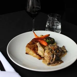 Dans cette image de Mariage Elegant, on voit un plat gastronomique servi au Doubletree par Hilton Québec Resort, ajoutant une note d'élégance pour une salle de réception de mariage. Le plat est composé d'un carré de viande avec une fine sauce aux champignons, accompagné de légumes frais, notamment des carottes et du brocoli. À côté de l'assiette, un verre de vin rouge et un verre d'eau complètent la présentation raffinée, avec en arrière-plan une nappe noire et des couverts élégamment disposés, reflétant une ambiance de réception sophistiquée.