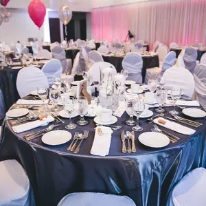 Dans cette image, la salle de réception du Doubletree par Hilton Québec Resort est magnifiquement aménagée pour un événement de mariage. Les tables rondes sont élégamment dressées avec des nappes blanches et des chemins de table gris foncé. Chaque table est décorée avec des compositions florales délicates au centre, entourées de verres à vin impeccablement disposés et de couverts en argent. Les chaises sont habillées de housses blanches, ajoutant une touche de sophistication. En arrière-plan, des rideaux et un éclairage rose créent une ambiance chaleureuse et romantique, tandis que des ballons flottent gracieusement dans un coin de la salle, contribuant à l’atmosphère festive. Cette image met en valeur la catégorie salle de réception pour Mariage Elegant, à Québec.
