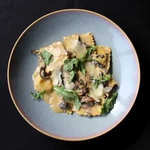 Le plat présenté est un ravioli garni de champignons, de roquette et de copeaux de parmesan, disposé artistiquement sur une assiette bleu-gris sur une nappe noire. Cette image est associée au Doubletree par Hilton Québec Resort, qui propose des s uperbes salles de réception parfaites pour les mariages.