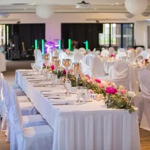 La photo montre une élégante salle de réception au Doubletree par Hilton Québec Resort, idéale pour les mariages. Des tables rectangulaires sont soigneusement dressées avec des nappes blanches impeccables et décorées de fleurs roses et blanches, ajoutant une touche de couleur et de romantisme. Des chaises recouvertes de housses blanches entourent les tables, contribuant à une atmosphère raffinée. En arrière-plan, un éclairage festif et des lumières colorées créent une ambiance joyeuse, parfaite pour célébrer des moments importants comme un mariage.