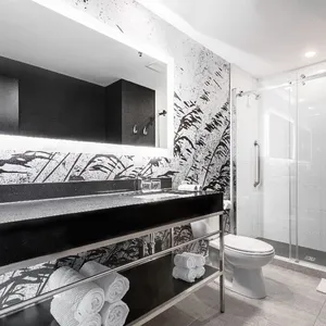 La salle de bain moderne de la salle de réception du Doubletree par Hilton Québec Resort présente un design élégant avec des murs ornés de motifs artistiques monochromes. Un large miroir rétroéclairé est suspendu au-dessus d'un comptoir en marbre sombre, agrémenté de plusieurs serviettes blanches soigneusement roulées. La douche à l'italienne est équipée de portes vitrées coulissantes, ajoutant une touche sophistiquée à l'espace. Ce cadre raffiné fait partie des installations proposées pour accueillir des événements de mariage mémorables.