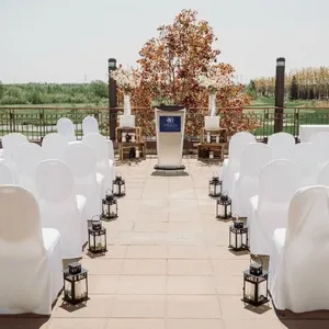 Dans cette image, la salle de réception extérieure du Doubletree par Hilton Québec Resort est préparée pour une cérémonie de mariage. Des chaises blanches soigneusement alignées sont disposées de chaque côté de l'allée centrale, menant à un podium orné du logo de l'hôtel. Chaque allée est bordée de petites lanternes noires, ajoutant une touche élégante et romantique à l'ambiance. En arrière-plan, un arbre décoratif et un paysage verdoyant complètent le cadre naturel et serein, idéal pour un mariage en plein air.