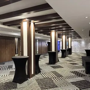 La salle de réception du Doubletree par Hilton Québec Resort est élégamment aménagée pour un événement de mariage. La pièce spacieuse est ornée de piliers en bois sombre et de plafond avec des panneaux lumineux. Le sol présente un motif géométrique en noir et blanc qui ajoute une touche moderne. Plusieurs tables hautes drapées de housses noires sont disposées pour accueillir les invités lors d'un cocktail. Cette salle de réception offre une ambiance raffinée et contemporaine, idéale pour célébrer un mariage mémorable.