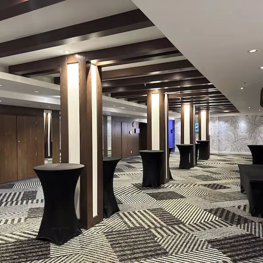 La salle de réception du Doubletree par Hilton Québec Resort est élégamment aménagée pour un événement de mariage. La pièce spacieuse est ornée de piliers en bois sombre et de plafond avec des panneaux lumineux. Le sol présente un motif géométrique en noir et blanc qui ajoute une touche moderne. Plusieurs tables hautes drapées de housses noires sont disposées pour accueillir les invités lors d