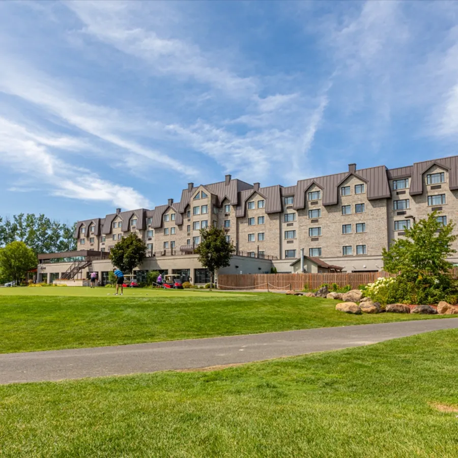 Le Doubletree par Hilton Québec Resort, une salle de réception prisée pour les mariages, est présenté ici sur une vaste pelouse verte sous un ciel bleu parsemé de nuages blancs. Le bâtiment est imposant avec une façade en pierre, plusieurs fenêtres et plusieurs étages. Des arbres encadrent le site, ajoutant une touche naturelle et élégante, idéale pour des cérémonies en plein air ou des réceptions raffinées.