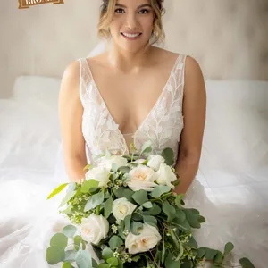 Cette image présente une mariée assise sur un lit, tenant un bouquet de fleurs blanches et vertes. La mariée porte une robe de mariée élégante avec un décolleté en V et des dentelles délicates. Ses cheveux sont coiffés en un chignon avec des mèches lâches encadrant son visage. Le style et l'élégance de la photographie mettent en avant le talent du photographe. PhotoMarc offre ses services dans la catégorie Photographie de mariage chez Mariage Elegant, Canada. Une impression de douceur et de joie se dégage de cette scène, capturant l’essence d’une journée de mariage parfaite.