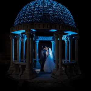 Dans cette image, un couple de mariés se tient sous un pavillon magnifiquement éclairé, avec une coupole ornée d'un éclairage bleu captivant. La mariée porte une robe blanche élégante et un long voile, tandis que le marié est vêtu d'un costume sombre. La scène crée une atmosphère romantique et magique, idéale pour immortaliser le moment. Cet exemple de photographie de mariage met en valeur le savoir-faire du prestataire PhotoMarc dans la capture d'instants mémorables et enchanteurs.