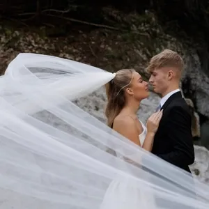 Dans l'image, un couple de jeunes mariés se tient face à face, entouré par un paysage naturel avec des rochers en arrière-plan et un petit ruisseau à leur côté. La mariée porte une robe blanche élégante avec un long voile léger qui se déploie gracieusement dans le vent, créant un effet spectaculaire. Le marié est vêtu d'un costume noir classique. Cette scène romantique et intime illustre le travail artistique de PhotoMarc dans la catégorie Photographie de mariage pour Mariage Elegant, Canada.