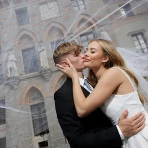 Un couple de mariés pose devant un château pittoresque avec une architecture superbe. La mariée, vêtue d'une élégante robe blanche, embrasse le marié, vêtu d'un costume noir classique. Le voile de la mariée s'envole gracieusement autour d'eux, ajoutant une touche romantique à la scène. Cette image illustre le service de photographie de mariage proposé par le prestataire PhotoMarc dans la catégorie photographie de mariage pour Mariage Elegant, Canada.