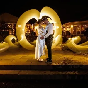 Un couple de mariés pose avec tendresse au centre d'une scène nocturne magnifiquement illuminée, capturée par PhotoMarc dans la catégorie Photographie de mariage. La mariée porte une robe blanche élégante, tandis que le marié est vêtu d'un costume sombre et élégant. Ils sont entourés de spectaculaires motifs lumineux dorés, ajoutant une touche de magie et de rêve à l'atmosphère. En arrière-plan, des bâtiments éclairés apportent une ambiance chaleureuse, parfaite pour célébrer un mariage.