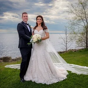 Un couple de jeunes mariés pose joyeusement devant un magnifique paysage lacustre sous un ciel partiellement nuageux. La mariée porte une élégante robe blanche ornée de dentelle, tenant un bouquet de fleurs blanches et vertes, tandis que le marié est habillé d'un costume noir classique avec un nœud papillon. Le couple se tient debout sur une pelouse verte avec des arbres en arrière-plan. Cette image, capturée par PhotoMarc, illustre la catégorie Photographie de mariage pour Mariage Elegant au Canada.