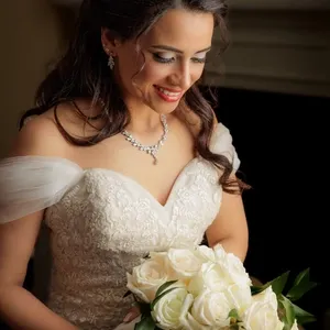 Une mariée souriante, vêtue d'une élégante robe en dentelle blanche, tient un bouquet de roses blanches. Ses cheveux sont coiffés en vagues douces et elle porte des bijoux scintillants qui complètent son allure raffinée. Cette image illustre parfaitement la catégorie Photographie de mariage, capturée par PhotoMarc pour Mariage Elegant, Canada.