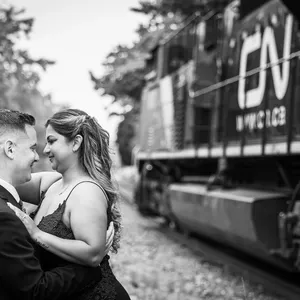 Un couple de jeunes mariés se tient proche l'un de l'autre dans un moment de tendresse sur fond de train en noir et blanc. La mariée porte une robe élégante et le marié, un costume chic. Le train, visible en arrière-plan, ajoute une touche unique et dynamique à cette scène romantique capturée par PhotoMarc, spécialiste en photographie de mariage. Ce cadre original est parfait pour illustrer l'alliance de romance et d'aventure, typique des souvenirs immortalisés par ce prestataire sur Mariage Elegant, Canada.