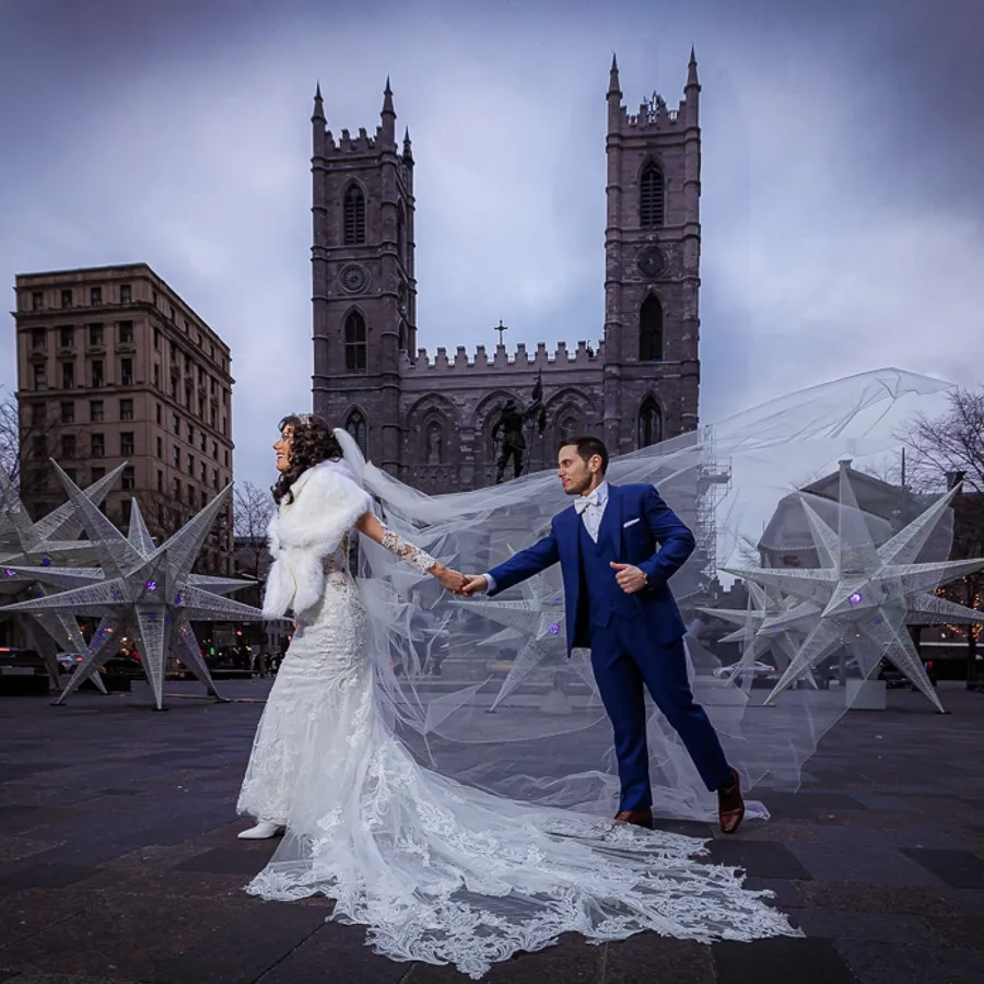 Un couple de mariés pose élégamment devant la Basilique Notre-Dame à Montréal, capturé par PhotoMarc pour la catégorie Photographie de mariage. La mariée porte une robe blanche ornée de dentelle avec une longue traîne et une étole en fourrure, tandis que le marié est en costume bleu. Ils se tiennent la main, entourés de décorations étoilées lumineuses, créant une atmosphère féerique idéale pour un mariage urbain chic.