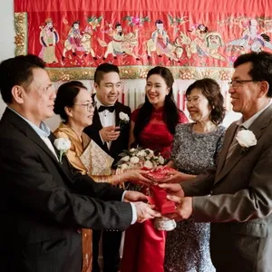 Dans cette image de Mariage Elegant, un groupe de six personnes se tient dans une ambiance festive, illustrant l'animation de mariage par KNL Productions. Deux couples plus âgés, vêtus de tenues élégantes, échangent des objets symboliques, illustrant une tradition culturelle. Deux jeunes mariés rayonnants, habillés d'une robe rouge pour la mariée et d'un costume sombre pour le marié, observent la scène avec des sourires éclatants. Derrière eux, une tapisserie rouge ornée de motifs détaillés ajoute une touche colorée et culturelle à la scène. Tous les participants montrent une grande complicité, symbolisant l'unité et la joie de ce moment de célébration.