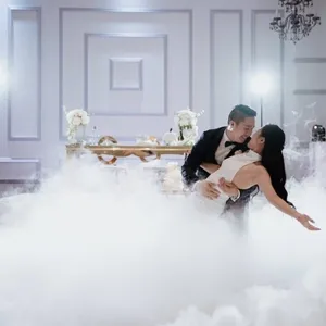 Dans cette image d'un mariage élégant au Canada, on voit un couple de jeunes mariés en pleine danse, entourés d'une brume mystique qui recouvre le sol, créant une atmosphère féerique. Les deux danseurs, vêtus d'une robe de mariée blanche et d'un costume sombre, s'embrassent dans une pose gracieuse. En arrière-plan, une table ornée de fleurs blanches apporte une touche de raffinement à la scène. Des lumières douces illuminent la salle, ajoutant à l'ambiance romantique de l'événement. La scène met en valeur les talents d'animation de KNL Productions, spécialisée dans la création d'ambiances mémorables pour les mariages.