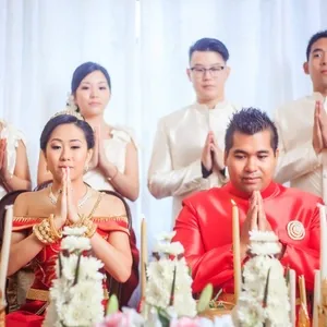 Mariage traditionnel où un couple, vêtu de tenues rouges ornées de bijoux dorés, est au centre de l'image, en train de prier ou de méditer lors de la cérémonie. Trois personnes se tiennent debout derrière eux, habillées en vêtements traditionnels blancs, également en prière. Le décor est agrémenté de fleurs délicates et d'éléments dorés, typiques des cérémonies de mariage. Cette scène est un exemple du travail de KNL Productions, spécialisé dans l'animation pour le mariage, sur le site de Mariage Elegant, Canada.