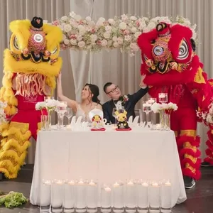 Dans cette image de Mariage Elegant, deux lions colorés de danse traditionnelle, l'un jaune et l'autre rouge, encadrent un couple de mariés assis à une table ornée de fleurs blanches et de bougies. La mariée porte une robe blanche élégante, tandis que le marié est en costume sombre. Autour de la table, des compositions florales et des bougies décorent l'espace, ajoutant une ambiance festive. Cette animation est fournie par KNL Productions dans le cadre de leur service d'animation de mariage.