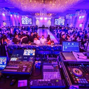 KNL Productions, expert en animation de mariages au Canada, présente une salle de réception somptueusement décorée pour un mariage. L'image montre une salle de bal élégante avec des tables rondes dressées, chacune ornée de nappes blanches et chaises stylées. Un éclairage bleu crée une atmosphère féerique, tandis qu'un plafond constellé de lustres brillants ajoute une touche sophistiquée. Au premier plan, une table de mixage et des équipements DJ, élément central du service d'animation de KNL Productions, sont prêts pour divertir les invités. Plusieurs écrans affichent des images, accentuant l’atmosphère festive de l’événement.