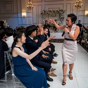 L'image montre une animatrice de mariage de KNL Productions dirigeant une activité interactive avec des invités assis, lors d'une réception de mariage élégante. Elle porte une robe élégante et tient un microphone, impliquant les invités qui sont habillés de manière formelle. L'arrière-plan présente un décor raffiné avec des chandeliers dorés et une belle composition florale sur un mur. Cet événement se déroule dans une salle somptueuse, parfaite pour une célébration de mariage, construisant une ambiance festive et chaleureuse qui met en valeur l'expertise en animation de KNL Productions dans le contexte des mariages.
