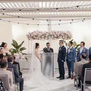 Dans cette image de la catégorie animation de Mariage Elegant, on voit la cérémonie de mariage se dérouler dans une salle lumineuse et contemporaine. L'espace est décoré avec raffinement, comprenant un grand arrangement de fleurs roses et blanches suspendues au-dessus de l'autel. La mariée et le marié, debout face à face, semblent échanger leurs vœux tandis que le célébrant les guide. À leurs côtés se trouvent les demoiselles d'honneur, habillées de robes élégantes, et les garçons d'honneur en costumes assortis, accompagnant le couple en cette occasion spéciale. Des invités sont assis, attentifs, créant une atmosphère intime et joyeuse. KNL Productions, expert en animation, assure l'ambiance et le bon déroulement de cet événement mémorable.