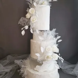 Ce magnifique gâteau de mariage de Patisserie Artisanale Kalina, catégorie Gâteaux de mariage, présente une élégante structure à trois étages. Chaque étage est recouvert d'un glaçage blanc immaculé avec une texture délicate. Des roses blanches soigneusement disposées et des feuilles délicates en sucre ornent le gâteau, ajoutant une touche de sophistication. Des éléments décoratifs translucides et aériens sont subtilement incorporés pour un effet moderne et artistique, parfait pour un mariage élégant et raffiné.