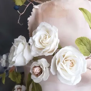 Gros plan sur un élégant gâteau de mariage créé par la Patisserie Artisanale Kalina, spécialisée dans les gâteaux de mariage. Le gâteau, d'un blanc immaculé, est orné de délicates fleurs en sucre magnifiquement réalisées, ressemblant à des roses blanches et des boutons de fleurs, accompagnées de feuilles vertes qui ajoutent une touche naturelle et raffinée. Les détails méticuleusement sculptés des fleurs apportent une texture riche et réaliste, capturant parfaitement l'artisanat et l'attention aux détails typiques des créations de la Patisserie Artisanale Kalina, idéale pour un mariage sophistiqué.