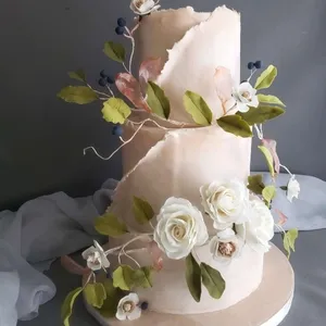 Ce magnifique gâteau de mariage à deux étages, créé par la Patisserie Artisanale Kalina, présente un design élégant avec une finition en crème blanche. Il est délicatement orné de roses blanches en sucre et de feuilles vertes, accentuées par des petites baies violettes et des motifs végétaux roses. Le thème naturel et romantique de ce gâteau s'accorde parfaitement avec des noces sophistiquées. L'ensemble repose sur un fond gris sobre, mettant en valeur les détails raffinés réalisés par ce talentueux fournisseur de gâteaux de mariage.