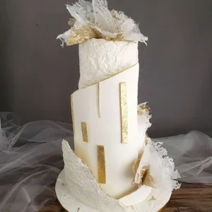 Cet élégant gâteau de mariage, réalisé par la Patisserie Artisanale Kalina, présente une structure verticale moderne. Il est recouvert d'un glaçage blanc texturé et orné de détails dorés rectangulaires qui ajoutent une touche sophistiquée. Des éléments en dentelle comestible blanche et dorée sont délicatement disposés sur le dessus et les côtés, accentuant le design artistique du gâteau. Idéale pour un mariage chic, cette pièce maîtresse incarne le savoir-faire artisanal et l'esthétique raffinée, correspondant parfaitement à la catégorie Gâteaux de mariage de Mariage Elegant, Canada.