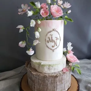 Gâteau de mariage élégant de la Patisserie Artisanale Kalina, présentant un design à trois étages. Le gâteau est décoré de délicates fleurs roses et blanches, soigneusement disposées autour du niveau supérieur, ajoutant une touche romantique. La base imite l'apparence du bois, apportant une ambiance rustique. Au centre, un monogramme est imprimé sur un fond blanc rehaussé de nuances rosées. Ce gâteau illustre l'expertise de la Patisserie Artisanale Kalina dans la catégorie des gâteaux de mariage, offrant une option visuellement époustouflante pour les couples à la recherche d'un design unique et sophistiqué.