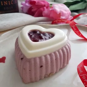 Un gâteau de mariage élégant de la Patisserie Artisanale Kalina est présenté ici, mettant en valeur un dessert en forme de cœur. Le gâteau est recouvert d'une mousse rose et garni au sommet d'un cœur de chocolat blanc rempli d'une confiture aux fruits rouges. À l'arrière-plan, des fleurs roses, un ruban rouge et une nappe en tissu blanc ajoutent une touche romantique, parfaite pour un mariage raffiné.