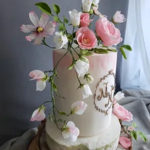 Gâteau de mariage élégant créé par Patisserie Artisanale Kalina, montrant une pièce montée cylindrique décorée de délicates fleurs roses et blanches en sucre. Le design inclut des détails floraux réalistes et un lettrage fin de couleur dorée sur la surface du gâteau, ajoutant une touche personnelle et raffinée. Idéal pour une cérémonie de mariage sophistiquée, ce gâteau de mariage se distingue par son esthétique naturelle et romantique, parfaitement adapté sur le site Mariage Elegant au Canada.