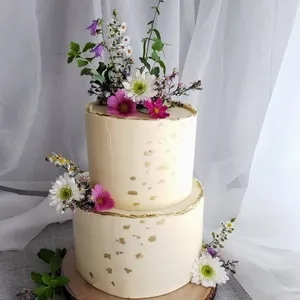 Gâteau de mariage à deux étages orné de fleurs fraîches, créé par Patisserie Artisanale Kalina, dans la catégorie des gâteaux de mariage. Le gâteau a une finition lisse avec des touches de dorure sur le glaçage blanc. Il est décoré de fleurs comme des marguerites blanches et des fleurs de couleur rose et violette, placées sur le dessus et entre les étages. Le gâteau repose sur un plateau en bois sur une nappe en tissu gris clair, apportant une touche naturelle et élégante idéale pour un mariage.