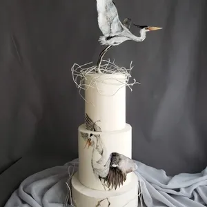 Gâteau de mariage élégant réalisé par Patisserie Artisanale Kalina, dans la catégorie Gâteaux de mariage. Ce gâteau à trois étages est minutieusement décoré, chaque étage arborant une couleur blanc cassé. Sur le dessus, une sculpture artistique d'un héron en plein vol est posée, ajoutant une touche de sophistication et de nature à la création. Les détails subtils de branches et de plumes attrayantes renforcent le thème naturel du gâteau. Une toile grise est drapée en arrière-plan, accentuant la simplicité et l'élégance du design.