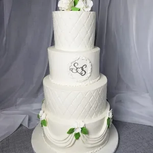 Le gâteau de mariage présenté par la Patisserie Artisanale Kalina, dans la catégorie Gâteaux de mariage sur Mariage Elegant, Canada, est un magnifique gâteau à trois étages. Chaque étage est délicatement recouvert de glaçage blanc, décoré de motifs en relief qui ressemblent à des losanges entrelacés. Le deuxième étage arbore un médaillon décoratif central portant des initiales élégantes. En bas de chaque étage, de petites fleurs blanches avec des feuilles vertes ajoutent une touche naturelle et raffinée. Au sommet, un bouquet de roses blanches complète l'ensemble, parfait pour un mariage élégant et sophistiqué.