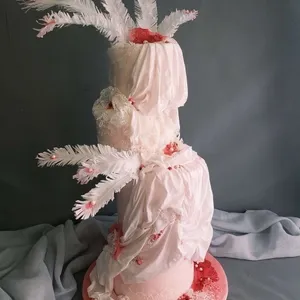 Gâteau de mariage artisanal créé par Patisserie Artisanale Kalina, catégorie Gâteaux de mariage. Ce gâteau élégant à trois étages est recouvert d'un drapé semblable à du tissu, ajoutant une touche sophistiquée. Des plumes blanches décorent le gâteau, accentuant sa hauteur et lui donnant une allure majestueuse. Des fleurs rouges et des détails délicats en dentelle ornent chaque niveau, créant un contraste harmonieux. Présenté sur une base rose pastel, ce gâteau est une pièce maîtresse idéale pour un mariage raffiné.