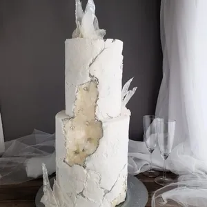 Gâteau de mariage à deux étages créé par Patisserie Artisanale Kalina, exposé sur la plateforme Mariage Elegant. Le gâteau est recouvert d'un glaçage blanc texturé, évoquant un aspect de pierre élégante. De délicats éclats de sucre translucides ornent le sommet et les côtés, ajoutant une touche artistique moderne. En arrière-plan, un tissu blanc drapé et deux flûtes à champagne en verre contribuent à l'ambiance raffinée de la présentation.