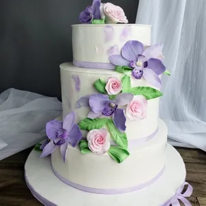 Gâteau de mariage à trois étages de la Patisserie Artisanale Kalina, décoré de fleurs en sucre violettes et roses, avec des touches de feuilles vertes. Le glaçage blanc lisse est orné de rubans mauves autour de chaque niveau. Le fond présente un rideau translucide laissant passer une douce lumière, créant une ambiance élégante. Ce gâteau, proposé par Mariage Elegant, est parfait pour un mariage sophistiqué au Canada.