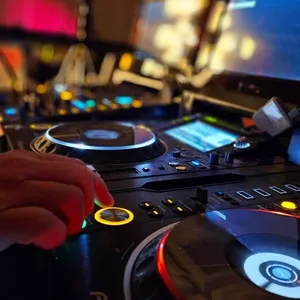 Dans l'image, on voit une table de mixage de DJ avec plusieurs platines et des lumières LED colorées, typiques de l'animation musicale lors de mariages. Une main ajuste les contrôles, illustrant l'engagement et le professionnalisme de Productions Megamix, un prestataire spécialisé dans l'animation de mariage. En arrière-plan, des écrans diffusent des lumières vives, ajoutant à l'ambiance festive qui est essentielle lors de célébrations de mariage à Mariage Elegant, Canada.
