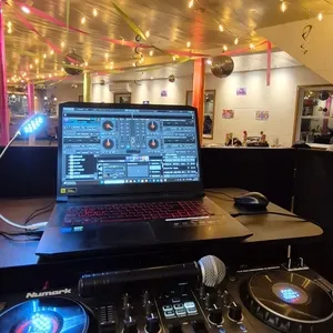 L'image présente une scène d'animation de mariage par Productions Megamix, dans un contexte de planification de mariage au Canada. Au premier plan, on peut voir une table de DJ équipée d'un contrôleur avec deux platines et un microphone, le tout placé sous un plafond décoré de guirlandes lumineuses et de boules à facettes. Un ordinateur portable affiche un logiciel de mixage musical, indiquant un environnement professionnel et festif. À l'arrière-plan, des invités sont assis à des tables, profitant de l'ambiance lumineuse et colorée, typique d'une réception de mariage animée.