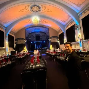 La photo présente une magnifique salle de réception ornée pour un mariage, illustrant les compétences de Productions Megamix dans la catégorie Animation. Deux longues tables sont élégamment dressées avec des nappes noires et des serviettes rouges, entourées de chaises noires. La salle est magnifiquement éclairée avec une lumière ambiante en dégradé de bleu et de rouge, créant une atmosphère festive. Au fond, des sapins illuminés ajoutent une touche de féérie. Au premier plan, un homme en costume noir sourit, ce qui reflète l'accueil chaleureux pour les invités. Cette scène démontre l'expertise de Productions Megamix dans la création d'ambiance pour des mariages mémorables en Canada.