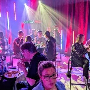 L'image montre un événement animé par Productions Megamix, spécialisé dans l'animation de mariages, avec des invités assis autour de tables hautes. La salle est élégamment éclairée par des lumières colorées, créant une atmosphère festive et joyeuse. Les participants semblent profiter de leur repas tout en discutant joyeusement. Le logo 'Megamix' est visiblement projeté sur le mur, soulignant la présence professionnelle du fournisseur dans le domaine de l'animation de mariages.