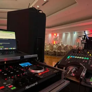 Dans cette image, on voit un équipement de son en préparation pour un événement de mariage, proposé par le prestataire Productions Megamix, spécialisé dans l'animation. À gauche, un ordinateur portable affiche un site web sur une table de mixage, tandis qu'à droite, une console de mixage avec divers contrôles lumineux est visible. En arrière-plan, une salle de réception élégamment décorée avec des chaises disposées autour de tables rondes et un éclairage d'ambiance rouge et blanc créent une atmosphère festive. Ce cadre met en valeur l'expertise de Productions Megamix dans la gestion de l'animation sonore pour des mariages sophistiqués au Canada.