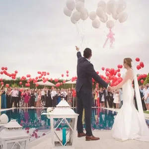 Un couple de mariés se tient au bord d'une piscine, main dans la main, regardant vers le ciel où des ballons blancs sont lâchés, symbole de célébration et d'un nouveau départ. Autour d'eux, une foule de convives, habillés avec élégance, tient des ballons rouges, ajoutant une touche vibrante à la scène. Des lanternes blanches décoratives sont posées au bord de la piscine, ajoutant une ambiance romantique et festive. Cette image représente un mariage à destination organisé par Voyage Club Evasion, dans le cadre magnifique d'une réception en plein air, typique de Mariage Elegant, Canada.