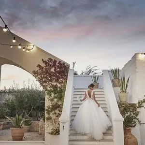 Une mariée en robe blanche élégante descend un escalier extérieur orné de plantes en pot sur le côté, sous un ciel au crépuscule. Autour, des guirlandes de lumières ajoutent une ambiance romantique. Le décor évoque une destination de mariage pittoresque. Ce cadre parfait est proposé par Voyage Club Evasion, expert en mariages à destination, présenté sur Mariage Elegant, Canada.