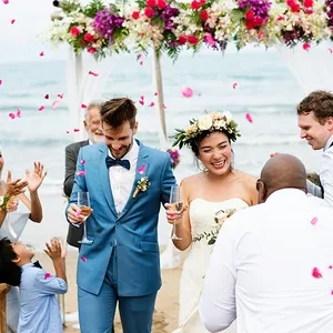 Un couple souriant vient de se marier sous une arche décorée de fleurs colorées, avec la mer en arrière-plan, lors d'un mariage à destination organisé par Voyage Club Evasion. La mariée porte une robe blanche élégante et une couronne de fleurs, tandis que le marié est vêtu d'un costume bleu. Les invités applaudissent et lancent des pétales de rose, créant une ambiance festive et joyeuse. Cette scène illustre parfaitement le service de mariage à destination offert par Voyage Club Evasion pour des célébrations inoubliables en bord de mer.