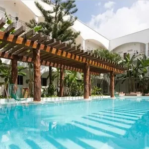 La photo montre une magnifique piscine extérieure entourée de chaises longues, située dans un cadre tropical avec des plantes exotiques et des palmiers. Un grand pergola en bois traverse la piscine, offrant une touche de charme rustique. En arrière-plan, le bâtiment de l'hôtel présente une architecture blanche et élégante avec des balustrades métalliques. Ce cadre paradisiaque est parfait pour un mariage à destination, catégorie proposée par le prestataire Voyage Club Evasion, spécialisé dans l'organisation de cérémonies de mariage exotiques et mémorables.